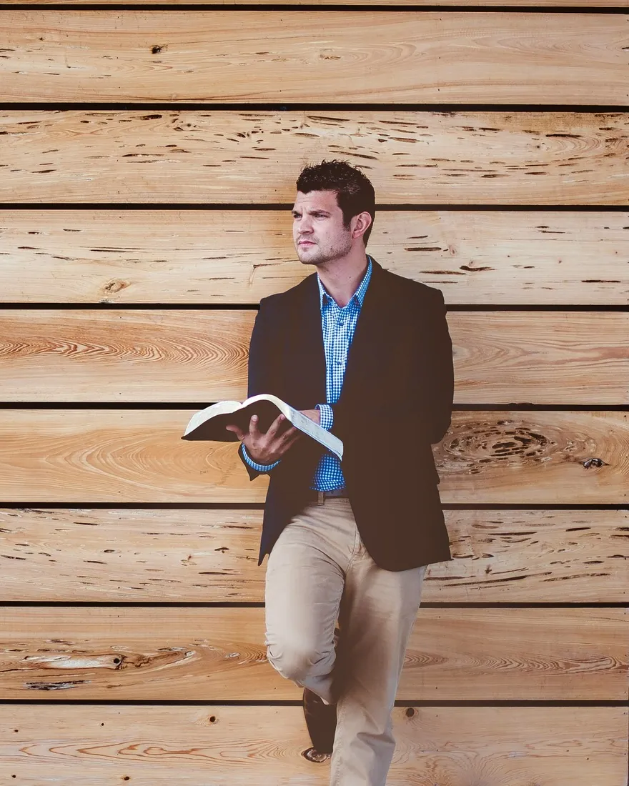 A man in a dark blazer leans against a wooden wall with an open book in his hands, looking off to one side.